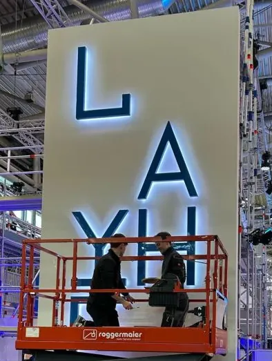 Zwei Monteure befestigen von der Hebebühne aus die Leuchtbuchstaben LAYHER an einer hohen Wand