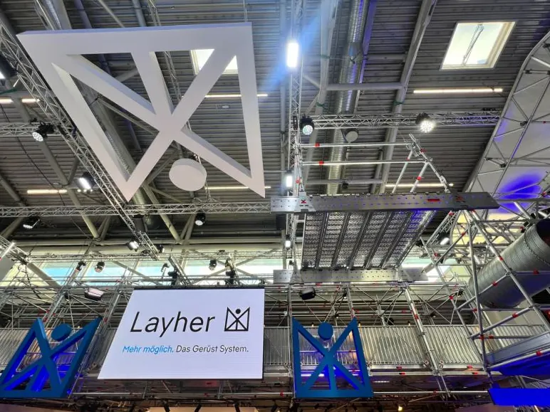 Blick unter die Decke des Layher-Stands: weißes Deckenelement, Gerüst und Bildschirm mit Layher-Logo
