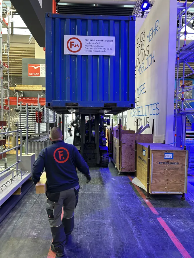 Ein Kollege von FREUNDE Messebau trägt Material in die Messehalle, hinter ihm bringt ein Stapler einen FREUNDE-Container